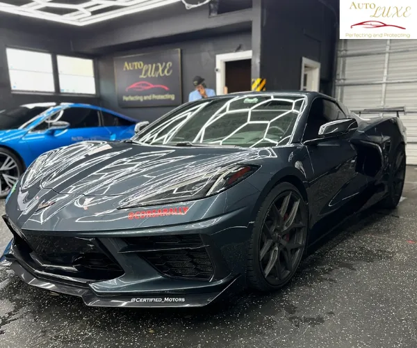 A dark gray high-performance sports car with a glossy finish parked in a professional detailing studio featuring overhead hexagonal lighting.