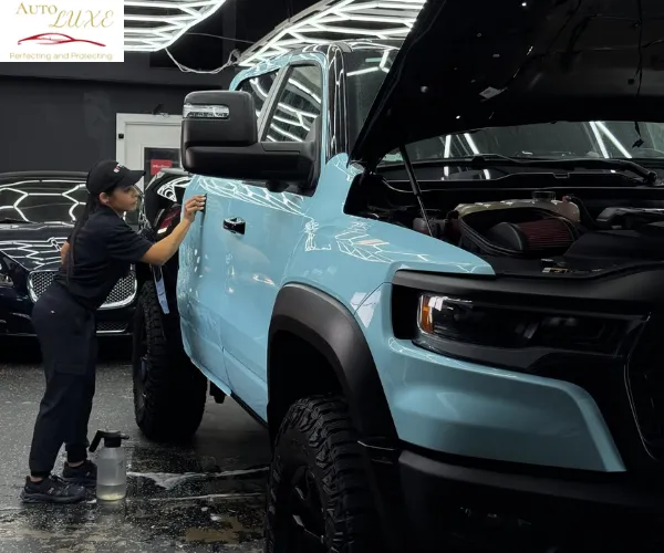 A professional technician applies a specialized coating to the door of a light blue performance truck with the hood open in a modern detailing studio.